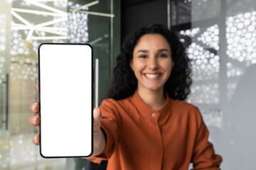 Successful business woman happily shows the smartphone screen to the camera, Latina woman with curly hair at the workplace recommends an online application.