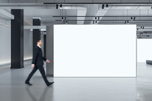Marketing Concept With Businessman Walking By Blank White Partition With Space For Advertising Poster Or Picture Frame In Loft Style Gallery Hall With Concrete Floor And Grey Wall Background, Mockup