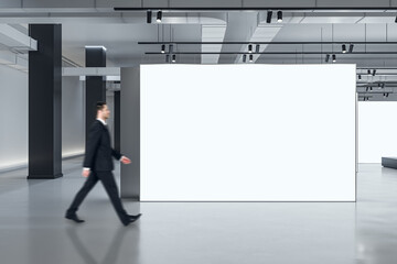 Marketing concept with businessman walking by blank white partition with space for advertising poster or picture frame in loft style gallery hall with concrete floor and grey wall background, mockup
