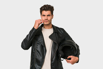 Young caucasian man holding a motrbike black helmet isolated with fingers on lips keeping a secret.