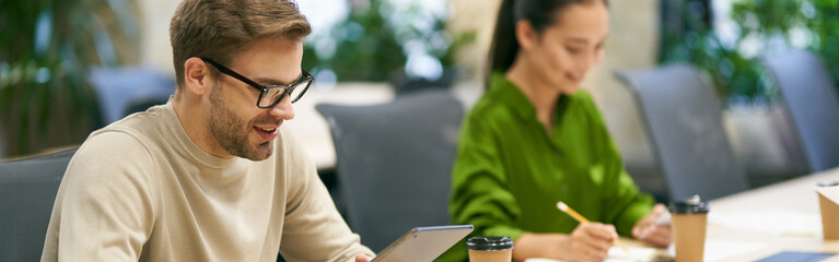 Wall Mural - Working together. Young smiling caucasian man wearing eyeglasses using digital tablet while sitting at desk with his female colleague in the modern coworking space