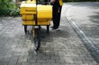 In schwarz-gelbgekleideter junger Postbote steht vor gelbem Fahrrad der Deutschen Post auf Gehweg in Stadt am Mittag im Frühling