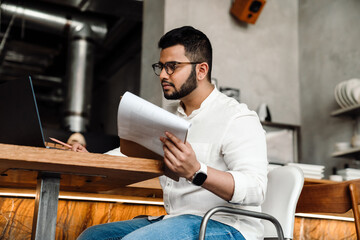 Side view of indian male business owner in eyeglasses filling orders on laptop in cafe