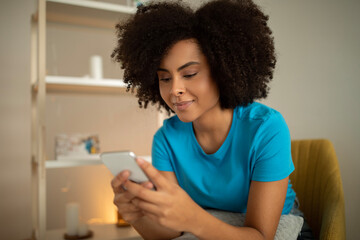 Glad millennial curly black lady typing on smartphone, watching video, surfing in internet