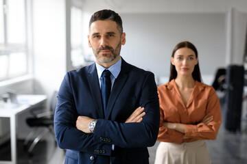 middle aged businessman and young businesswoman with arms crossed posing in office and looking at ca