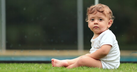 Adorable baby in outdoor grass garden backyard looking at children play inside pool