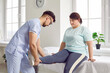 Physiotherapist examines overweight female patient before offering treatment therapy. Doctor doing manipulations with leg of fat, plus size young woman sitting on medical bed at physiotherapy clinic