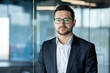 © Tetiana - Portrait of a young male student, intern seriously looking at the camera, standing in an office, campus, co-working space. Passes an interview, studies, takes an exam.