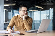 © Tetiana - Tired and bored young man businessman, accountant, manager sitting at a table and working on a laptop and with documents. Propping his head with his hand, he looks disappointedly at the monitor