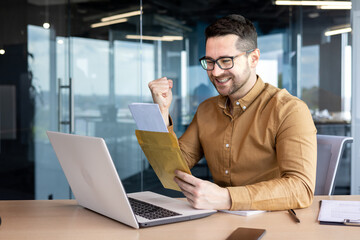 Happy young man businessman opens an envelope with a letter at the table in the office. Received good news, rejoices, shows a victory gesture with his hand