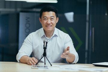 Asian smiling young man sitting in office at desk in front of camera, talking on microphone via video call, blogging, webinar, training, recording podcast.