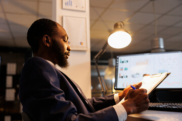 Careful entrepreneur analyzing marketing graphs, working late at night at company investment plan in startup office. African american worker planning strategy to help increase profit