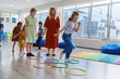 © .shock - Small nursery school children with female teacher on floor indoors in classroom, doing exercise. Jumping over hula hoop circles track on the floor.