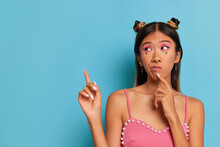 Thai Girl With Funny Hairstyle In Pink Top Posing Against Blue Wall, Presses Forefinger To Her Chin And Pointing Another Finger Up, Good Offer Concept, Copy Space