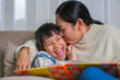 © Pornpimon - Mother reading a book with her daughter on the sofa at home. Happy mother reading a book, relaxing, hugging, telling funny stories to children and spending time together at home.