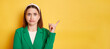 © sementsova321 - Confused puzzled woman wearing green jacket posing isolated over yellow background pointing away at copy space, advertisement area mockup.