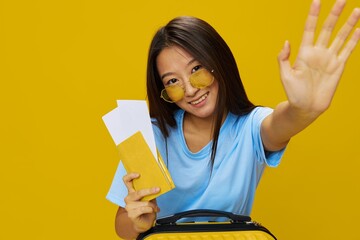 Wall Mural - Asian woman traveling with yellow suitcase and tickets with passport in hand, tourist traveling by plane and train with luggage on yellow background in blue T-shirt and jeans