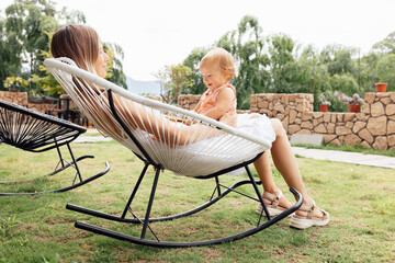 Wall Mural - Portrait of young Caucasian mother holds hug little toddler baby one year old, relax together outdoor on backyard at hot summer day. Happy mom embracing small infant child showing love and care