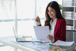 © PHAISITSAWAN - Asian businesswoman in red suit calculating company sales with calculator, laptop, and tablet on table Interior of businesswoman office at the modern workstation.