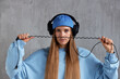 Portrait of a young pretty long-haired girl in a blue sweater and cap wears DJ headphones on her head and holds a wire in her hands. Music, fun and beauty.