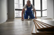 © anatoliy_gleb - Man in work overalls installing laminate timber flooring in living room with large window. Male construction worker laying laminate wooden floor in apartment under renovation.