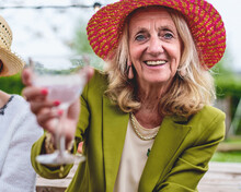 Blonde Senior Woman In Red Straw Hat Toasting At A Table With Friends, Invitingly Smiling At The Camera.