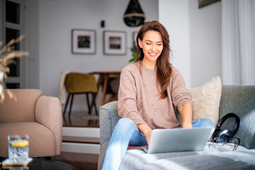 Wall Mural - Smiling caucasian woman sitting on the sofa at home and using laptop