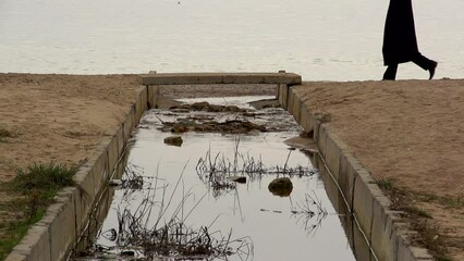 Wall Mural - A young guy in a black coat crosses the river on a small bridge against the background of the sky and the sea