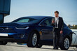 © standret - Serious facial expression. Businessman is standing near his electric car outdoors