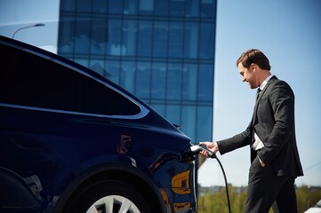 Man in suit and tie is holding charger and standing near his electric car outdoors