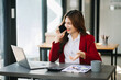 © Nuttapong punna - Business asian woman Talking on the phone and using a laptop with a smile while sitting at modern office.