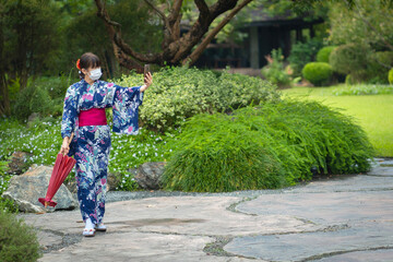 Cute Japanese women wearing beautiful traditional clothes dress walking relax happily in casual Yukata kimonos with white masks and red umbrellas in a fresh green natural garden.