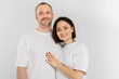 cheerful and tattooed woman with short brunette hair hugging joyous husband with bristle while standing together in white t-shirts and looking at camera isolated on grey background, happy couple