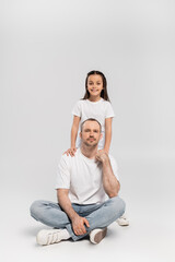 Wall Mural - happy preteen girl with brunette hair hugging cheerful father with bristle while posing in white t-shirts and blue denim jeans and looking at camera on grey background, Happy Father`s Day