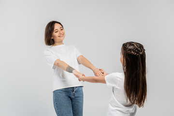 Wall Mural - happy mother with tattoo on hand and short brunette hair holding hands with preteen daughter while standing together in white t-shirts and blue denim jeans on grey background, child protection day