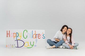 Wall Mural - Full length of smiling mother in casual clothes hugging preteen daughter and looking at camera together near placard with happy children's day lettering on grey background