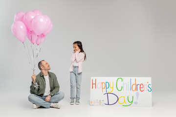 Wall Mural - Excited preteen girl in casual clothes looking at smiling father holding festive pink balloons near placard with happy children's day lettering during celebration on grey background