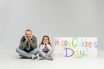 Wall Mural - Smiling father and preteen daughter in casual clothes looking at camera while sitting near placard with happy children's day lettering during celebration in June on grey background