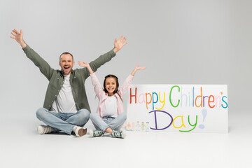 Wall Mural - Excited and cheerful man and preteen daughter in casual clothes looking at camera together while sitting near placard with happy children's day lettering on grey background