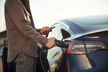 Grey colored formal clothes. Man is standing near his electric car outdoors