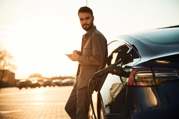 Leaning on the automobile and holding smartphone. On the charging station. Man is standing near his electric car