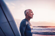 © ADDICTIVE STOCK - Generative AI image of side view of bald middle aged male in blue wetsuit standing near surfboard and looking away against cloudy sundown sky and wavy seafront