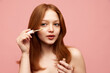 © master1305 - Portrait of young, beautiful, redhead girl with freckled skin applying concealer on face against pink studio background. Concept of natural beauty, makeup, cosmetology, cosmetics, skin care