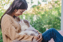 Woman With Long Hair In Brown Sweater And Jean Holding Persain Cat In Her Lap And Embrassed Him With Love Feelling, Girl And Cat Looking Each Other With Love And Care Emotion.