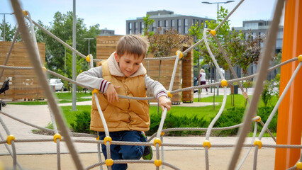 Wall Mural - Cheerful boy climbing through ropes and obstacles on playground at modern living block outdoors