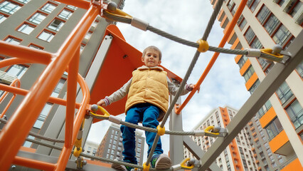 Wall Mural - View from the ground on little boy walking over the rope bridge and nets at playground. Active child, sports and development, kids playing outdoors