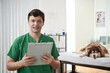 © DragonImages - Portrait of happy veterinarian with clipboard standing in office cleanic, samoyed dog in background