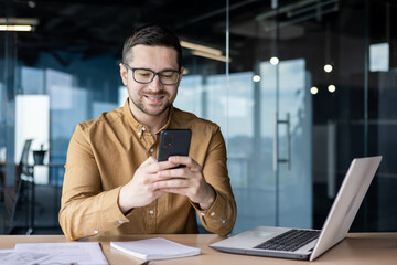 smiling young male office worker sits at a desk with a laptop and writes a message on the phone, cha