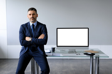 Poster - Confident middle aged businessman leaning on table, posing with folded arms near computer with blank screen, mockup