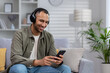 © Tetiana - Young hispanic man at home sitting on sofa resting, man with headphones listening online music, radio and audio books podcasts in app on phone